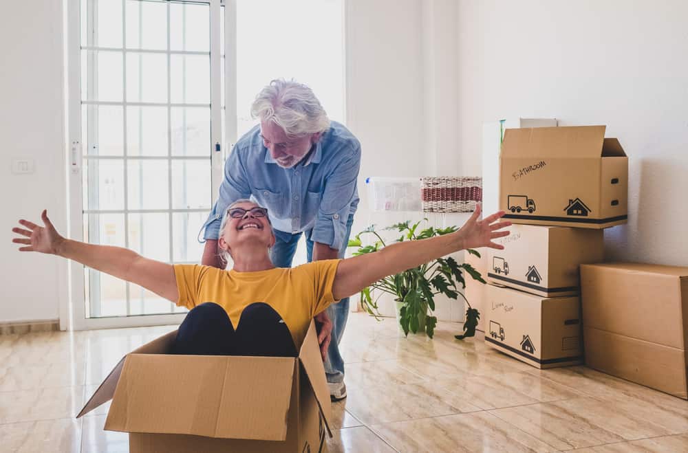 A senior woman joyfully sits in a moving box as a senior man pushes her